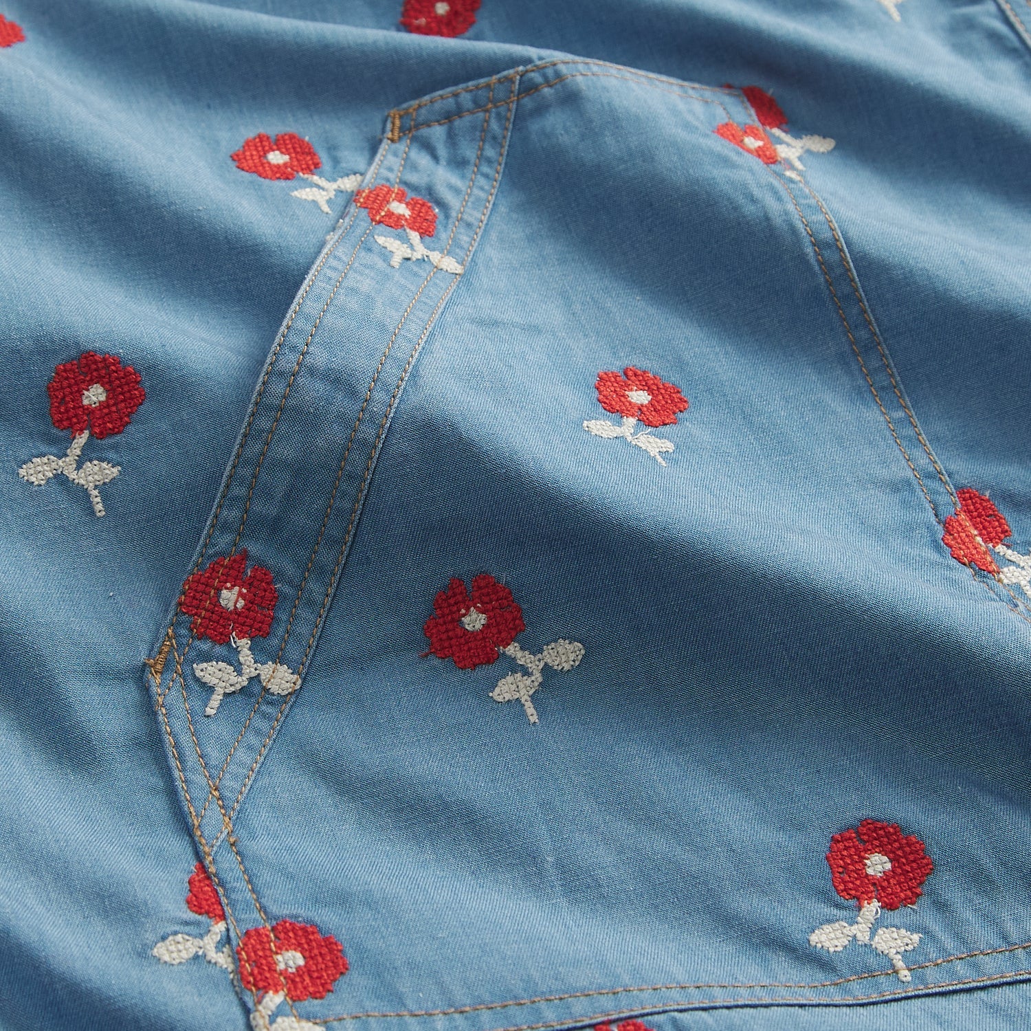 Close-up flat detail of light blue denim robe pocket area with red cross-stitch poppy flower embroidery