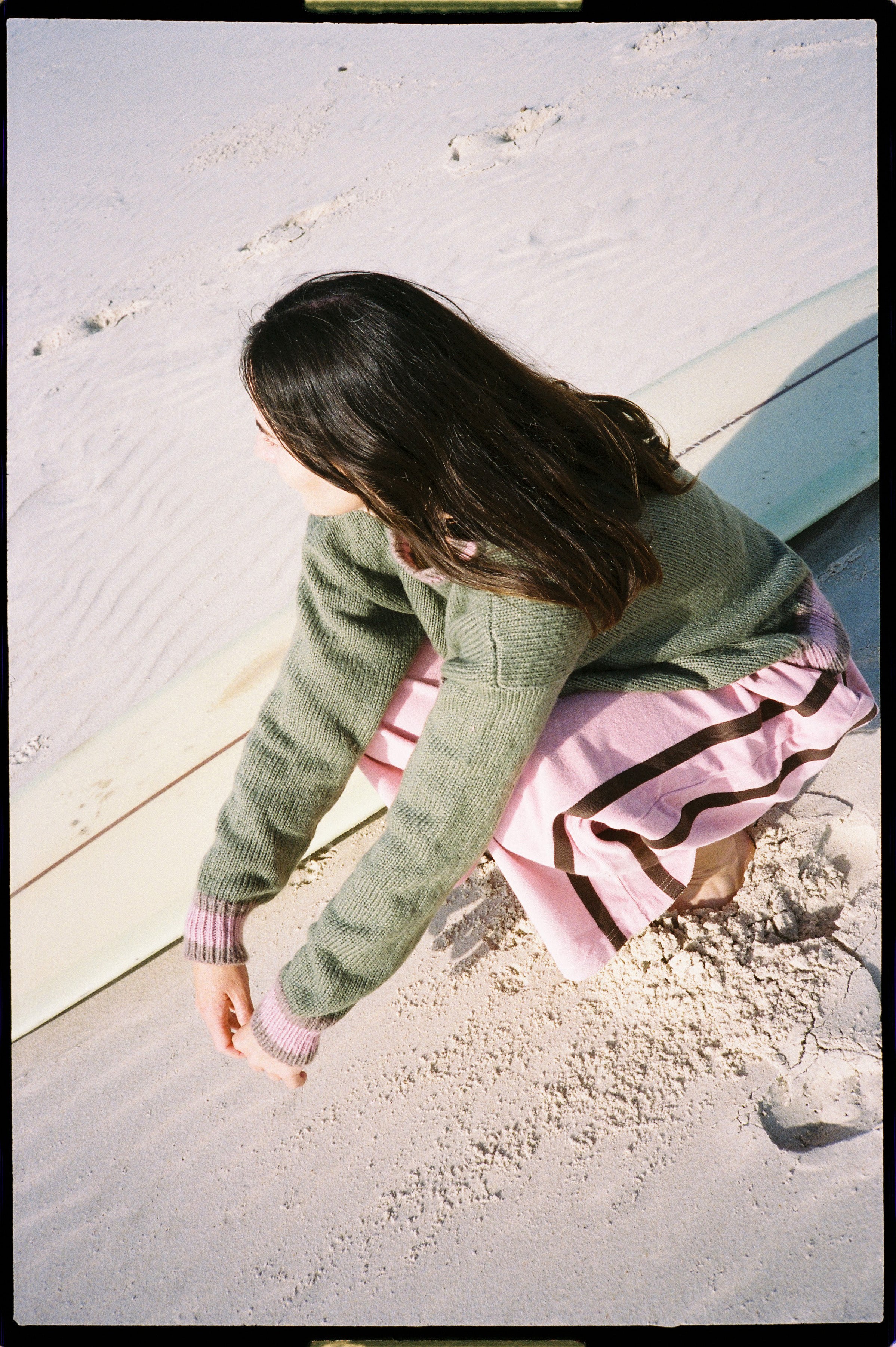 Overhead angle of model in blush pink Après Surf Panel Pants sitting on beach with surfboard