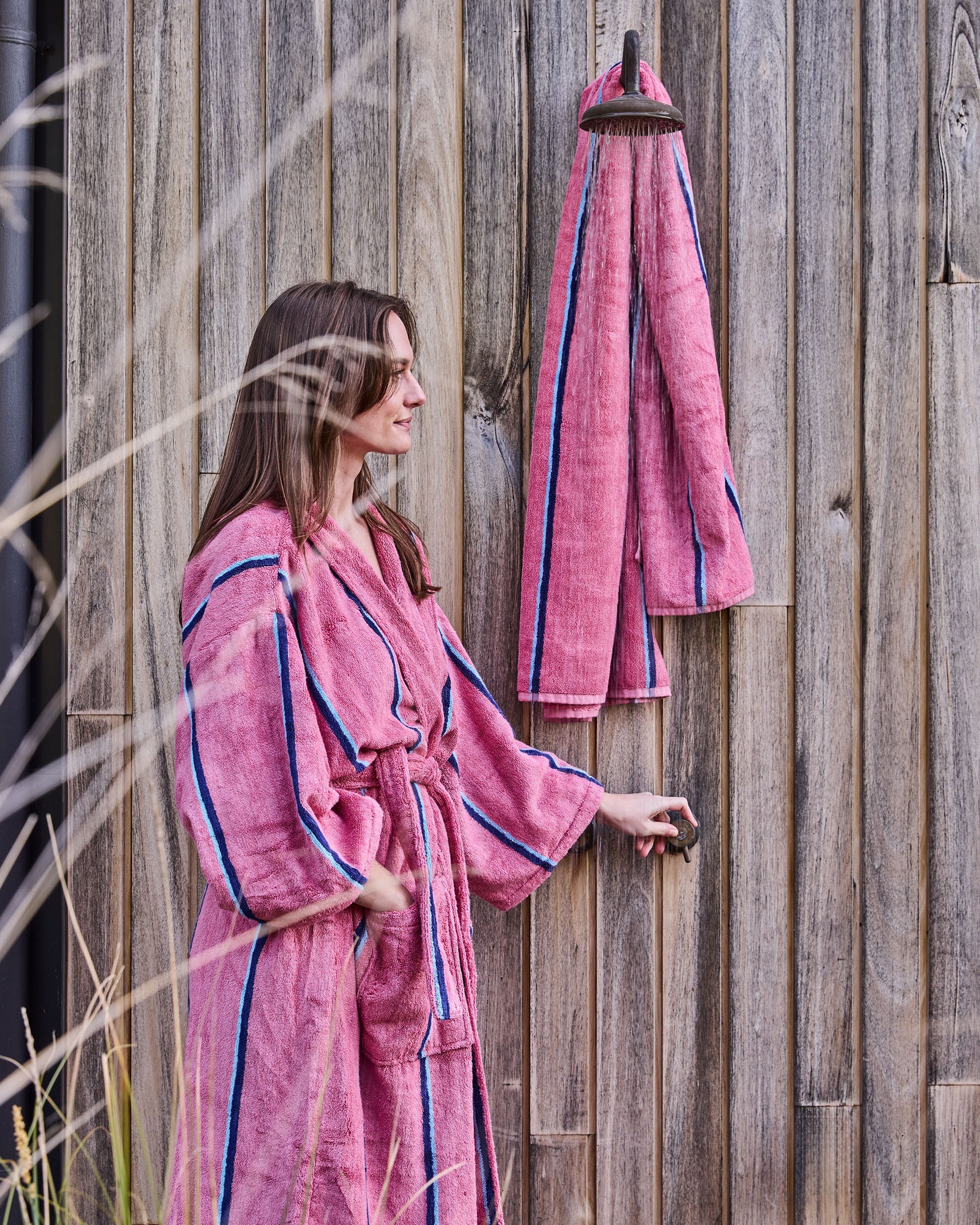 Outdoor lifestyle shot of model in matching Blushing Stripe robe with bath sheet hanging on wooden fence