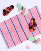 Overhead view of pink Kip Co Blushing Stripe Bath Mat with blue stripes styled with colorful slippers