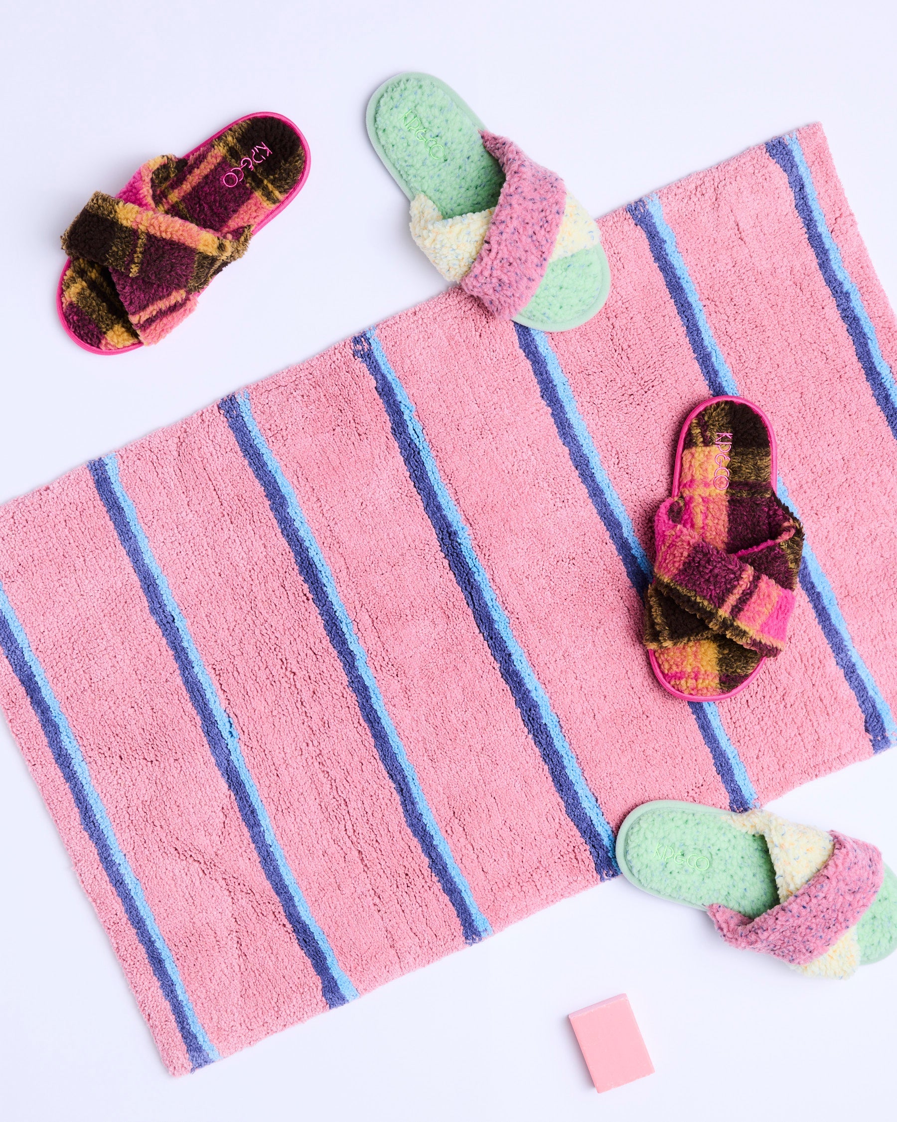 Overhead view of pink Kip Co Blushing Stripe Bath Mat with blue stripes styled with colorful slippers