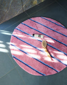 Top view of Kip & Co Blushing Stripe Round Bath Mat on dark tile floor with bath accessories and brush