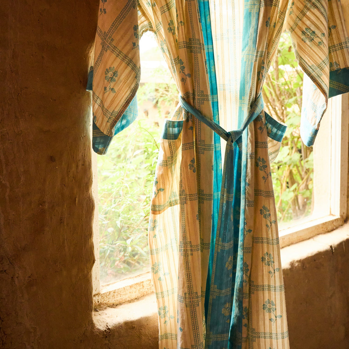 Turquoise Bonny Cotton Robe hung by a window, showing floral check print and turquoise belt and trim detail