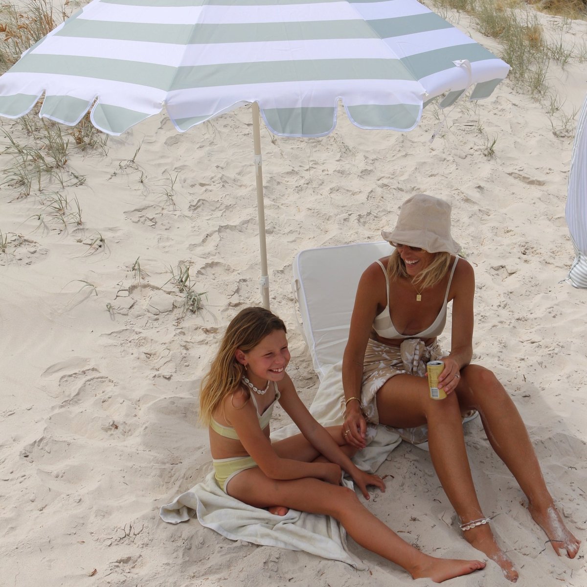 Sea sage green beach umbrella tilted on sandy beach providing shade for two people lounging underneath