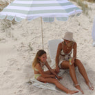 Sea sage green beach umbrella tilted on sandy beach providing shade for two people lounging underneath