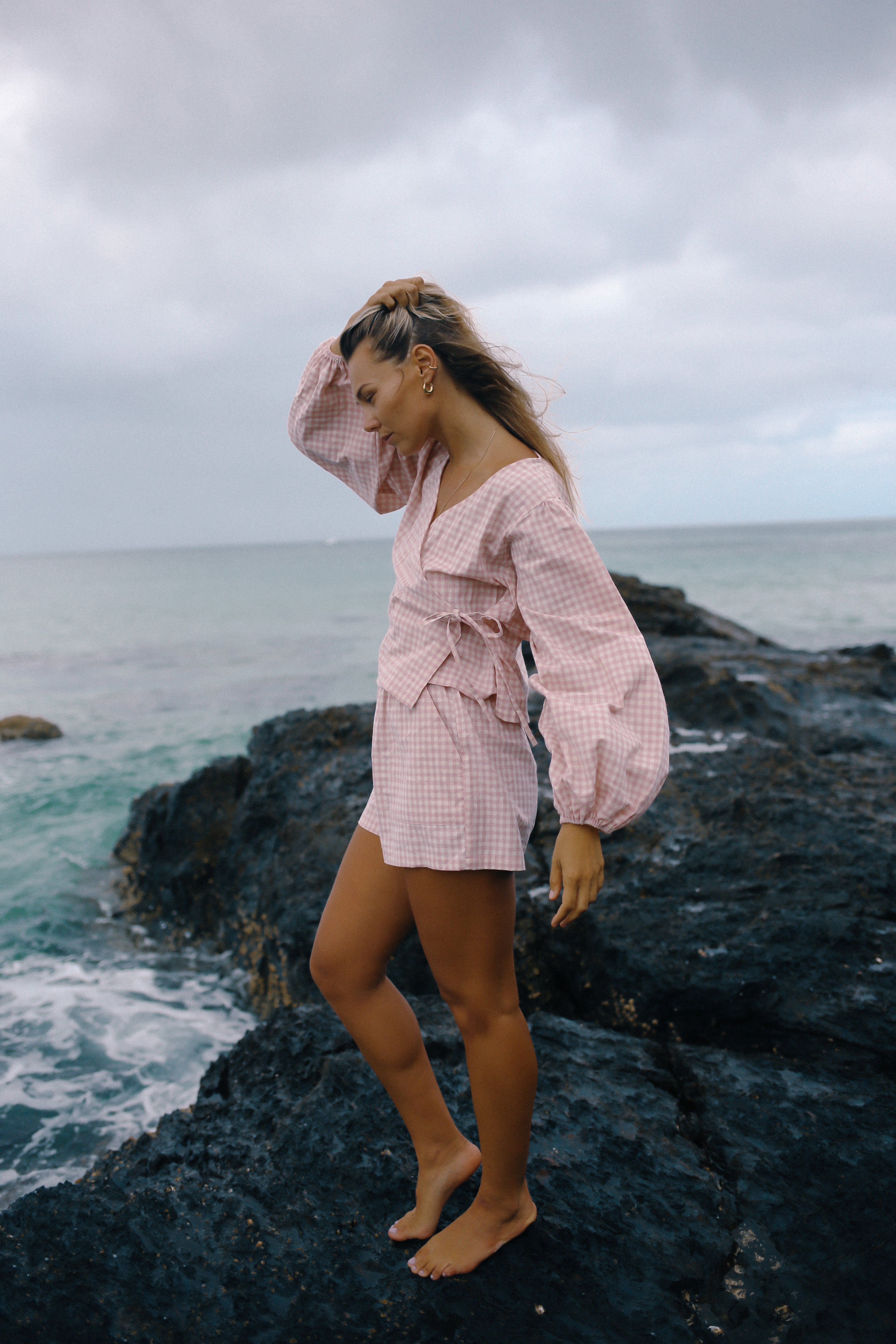 Woman in a pink outfit standing on a rocky outcrop by the ocean under a cloudy sky. Ella Wrap Top pink gingham by The Bare Road