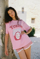 Woman wearing a pink 'Fraise' t-shirt and checkered shorts leaning against a tree