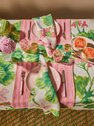 Colorful Bonnie & Neil table setting with floral tablecloth, pink plates, and watermelon on a woven surface.