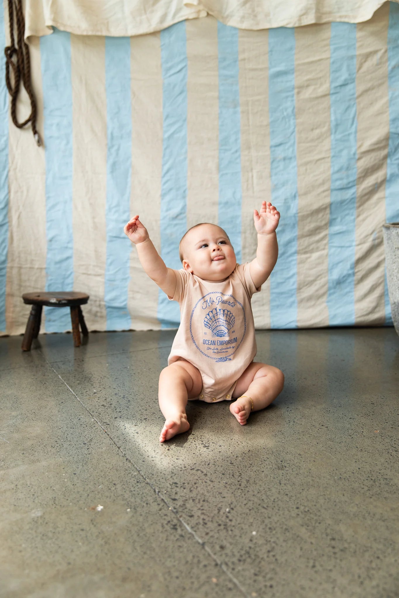 Baby wearing cream colours onesie with blue shell graphic "Miss Pearls Ocean Emporium" by Pearson & Pearson