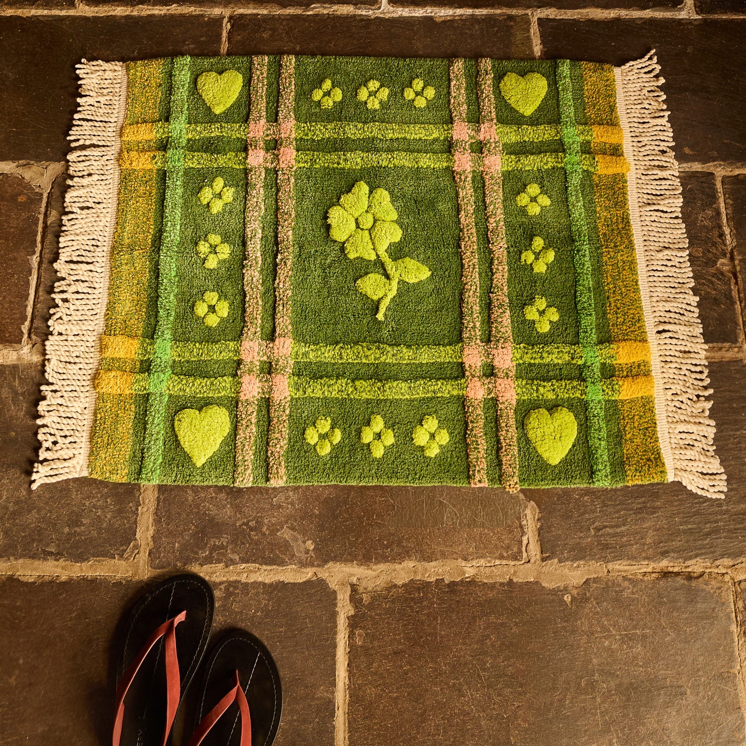 Green tufted bath mat with heart and floral motifs laid on stone floor tiles, viewed from above