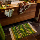 Green tufted bath mat with plaid and floral design styled on a dark tile bathroom floor beside a wooden vanity