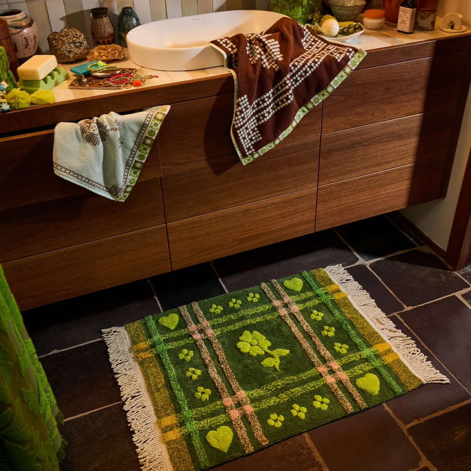 Green tufted bath mat with plaid and floral design styled on a dark tile bathroom floor beside a wooden vanity