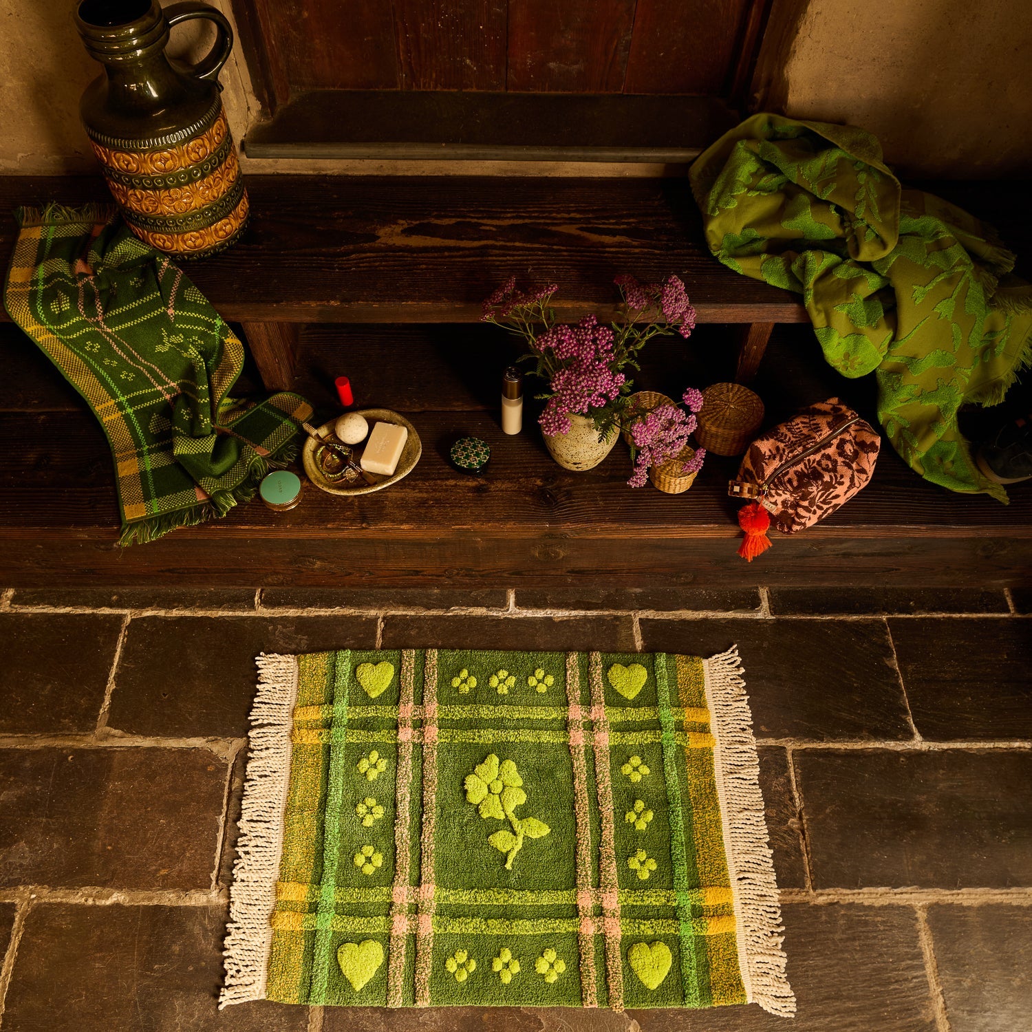Green plaid tufted bath mat with heart and flower motifs and cream fringe, laid on stone floor from above