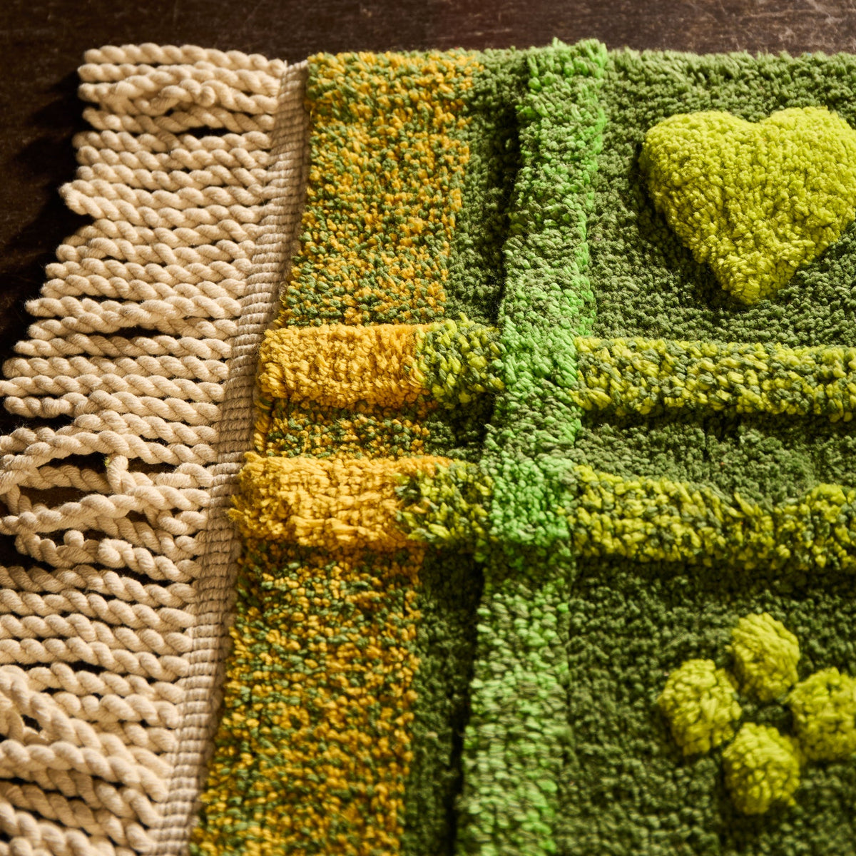 Close-up of green tufted bath mat showing cream twisted fringe, yellow plaid border, and raised heart motif texture
