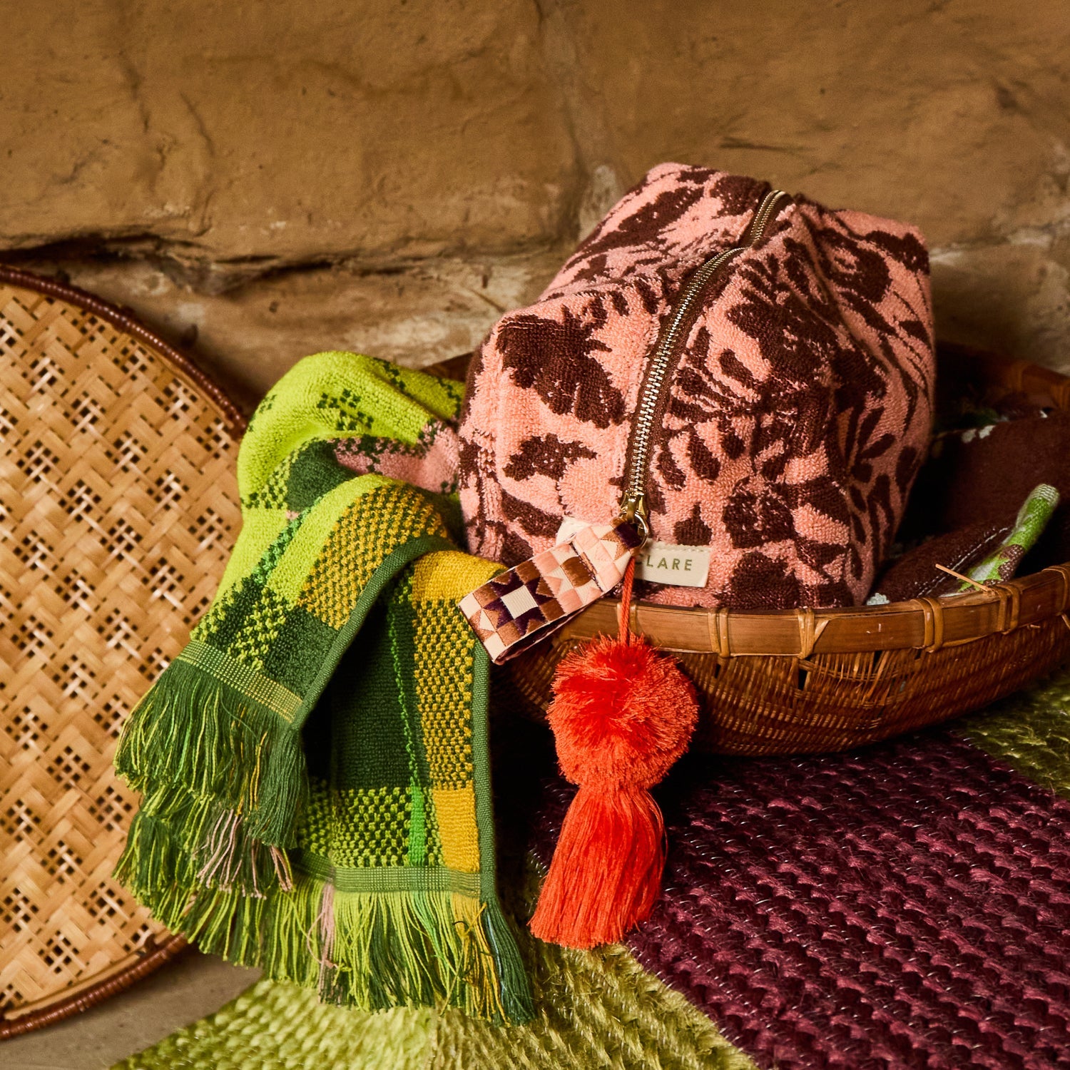 Green plaid fringed hand towel styled with a pink floral toiletry bag and wicker basket in a rustic setting