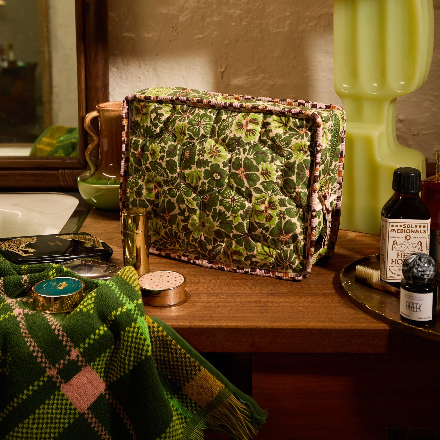 Green plaid fringed hand towel in the corner of a styled vanity scene with a floral quilted toiletry bag