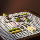 Collection of cheese knives with colorful twisted handles displayed on tiled surface with fruit and vegetables