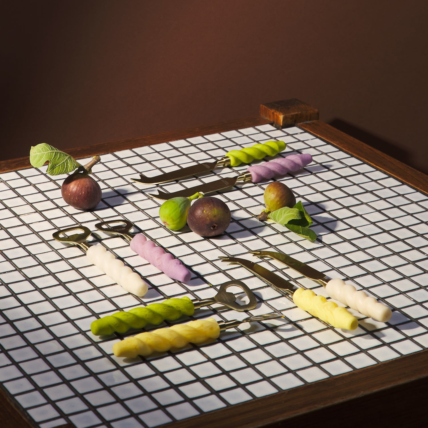 Collection of cheese knives with colorful twisted handles displayed on tiled surface with fruit and vegetables