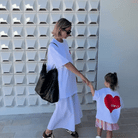 Woman in a white outfit holding hands with a child wearing a shirt with a red heart design, against a geometric wall.