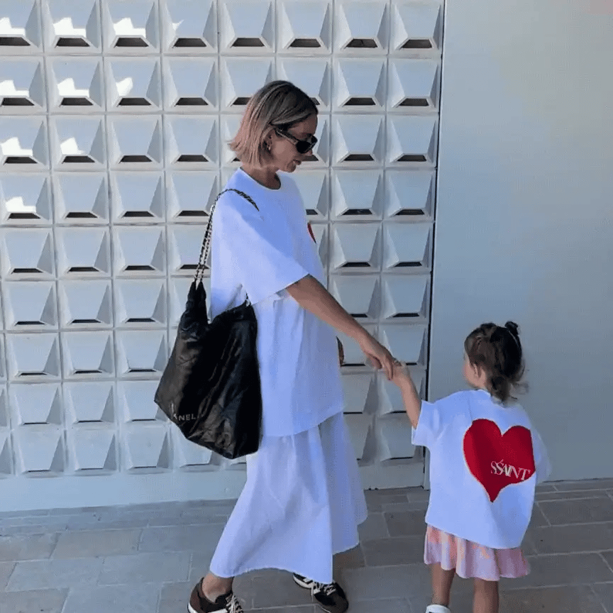 Woman in a white outfit holding hands with a child wearing a shirt with a red heart design, against a geometric wall.