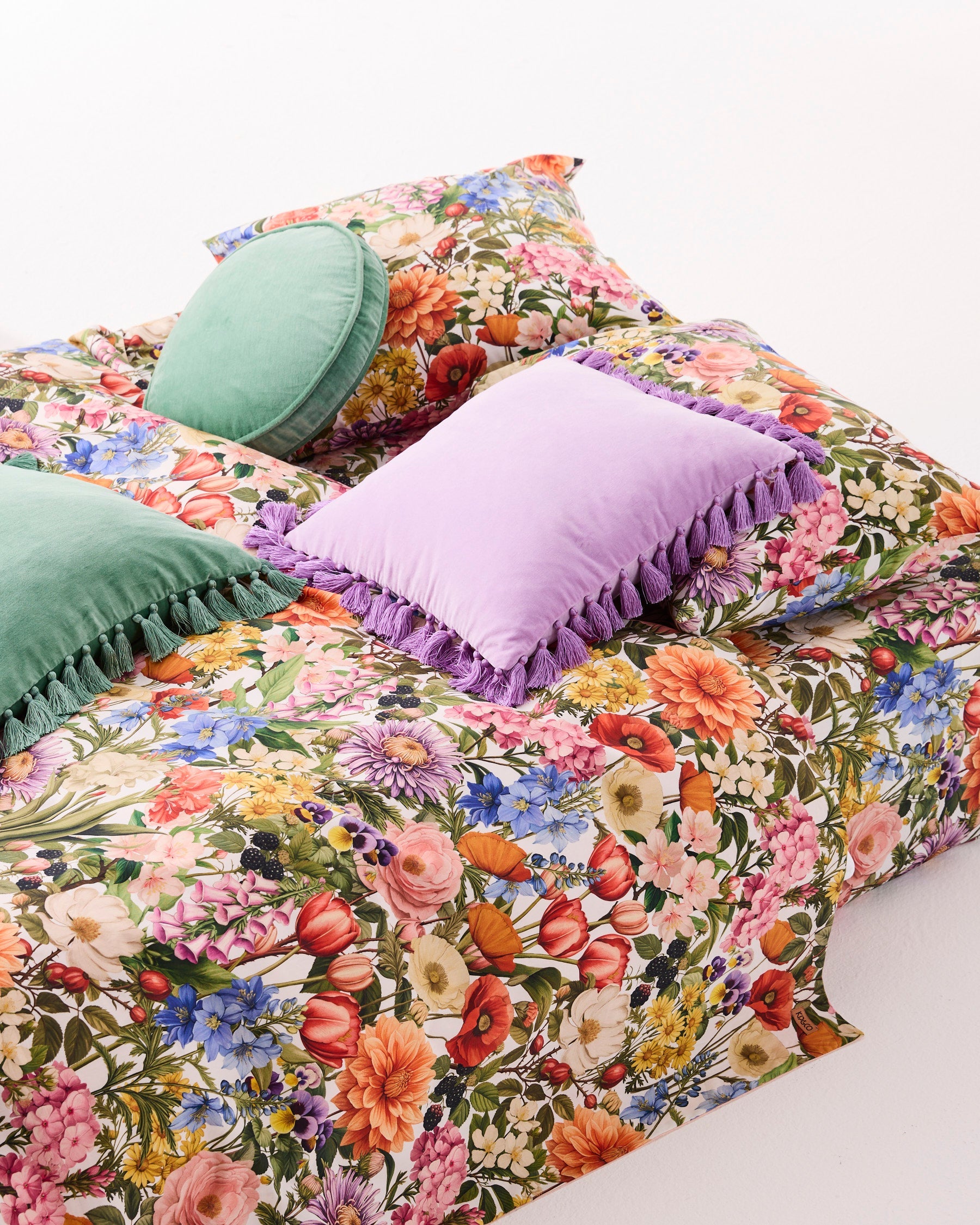 Lifestyle shot of lilac velvet tassel cushion styled with floral bedding and green velvet cushions