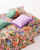 Lifestyle shot of lilac velvet tassel cushion styled with floral bedding and green velvet cushions