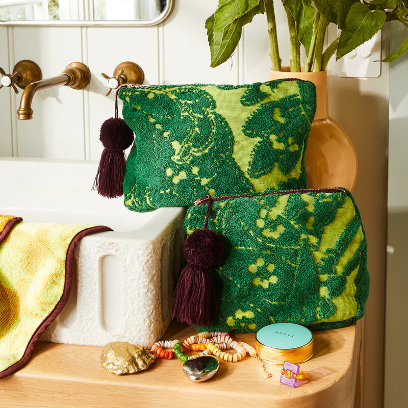 Green patterned pouch with burgundy tassels on a wooden surface next to a plant and sink.
