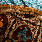 Close-up of brown velvet bedcover surface showing teal floral motifs within ornate medallion frames and gold fringe trim