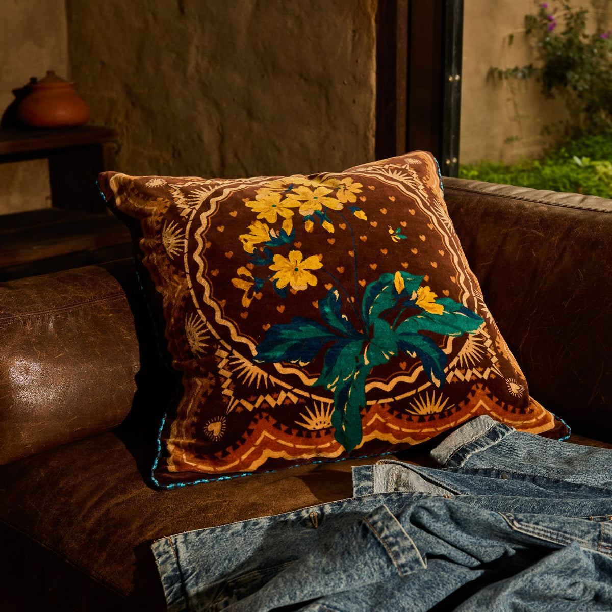 Brown velvet cushion with yellow floral and teal leaf print propped on a leather sofa in a warm interior setting