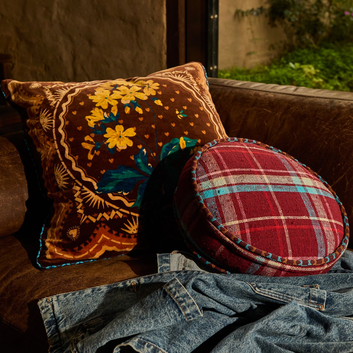 Brown floral velvet cushion styled on a leather sofa alongside a round red tartan cushion
