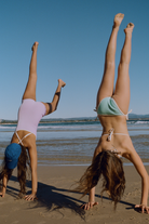 Two women doing cartwheels on the beach, one wearing a lilac ribbed onesie with white crossback straps