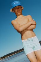 Boy wearing aqua mini stripe surf shorts on the beach, arms crossed, low-angle shot against blue sky