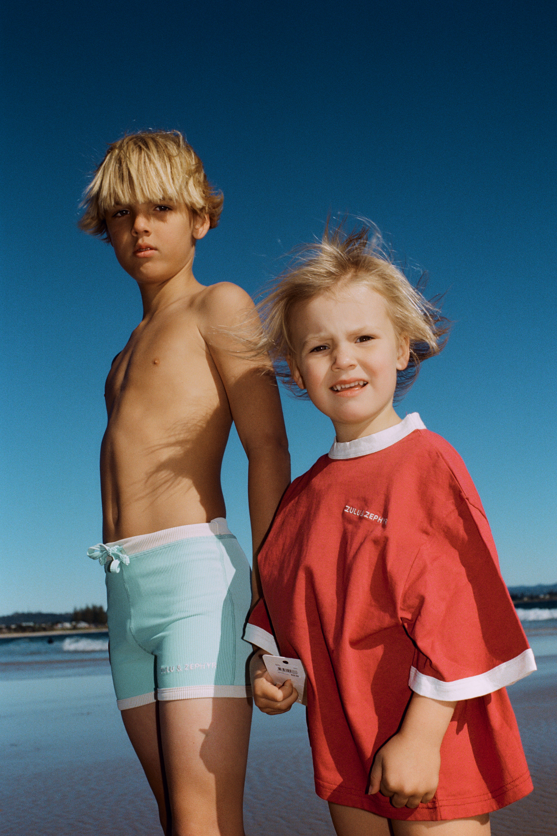 Two children on the beach, boy wearing aqua mini stripe surf shorts alongside a girl in a red top