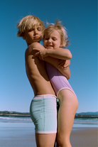 Two children hugging on the beach, boy wearing aqua mini stripe surf shorts with white contrast trim