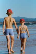 Two children walking on the beach wearing blue mini stripe surf shorts and matching swimwear, rear view