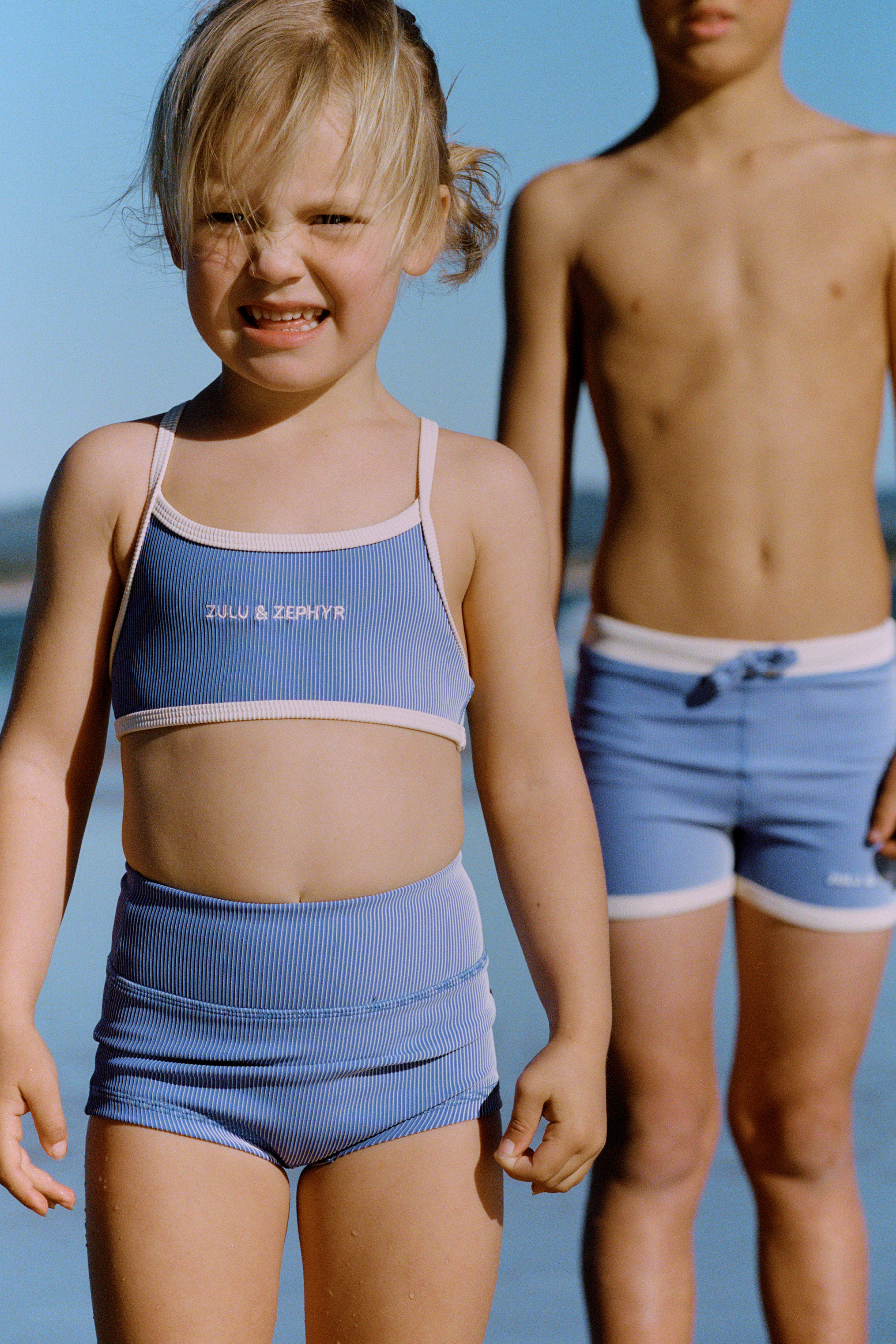 Child at the beach wearing blue mini stripe surf shorts with white trim, shown on a boy in the background