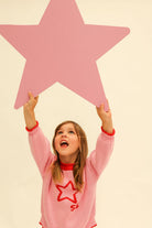 Child wearing pink Neve Knit sweater with red star motif reaching up toward large pink star prop