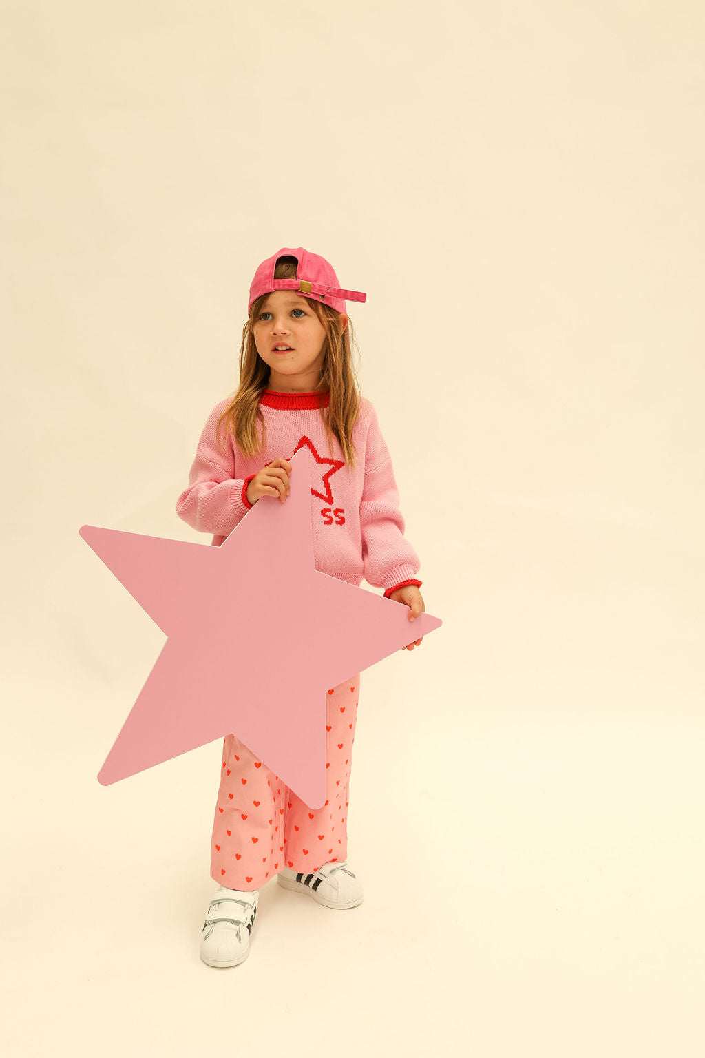 Full body shot of child in pink Neve Knit sweater with red star design and pink baseball cap