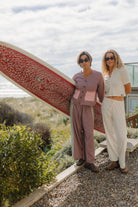 Two women modeling Atmosea Patchwork Pant Houndstooth Combo in coastal setting with surfboard