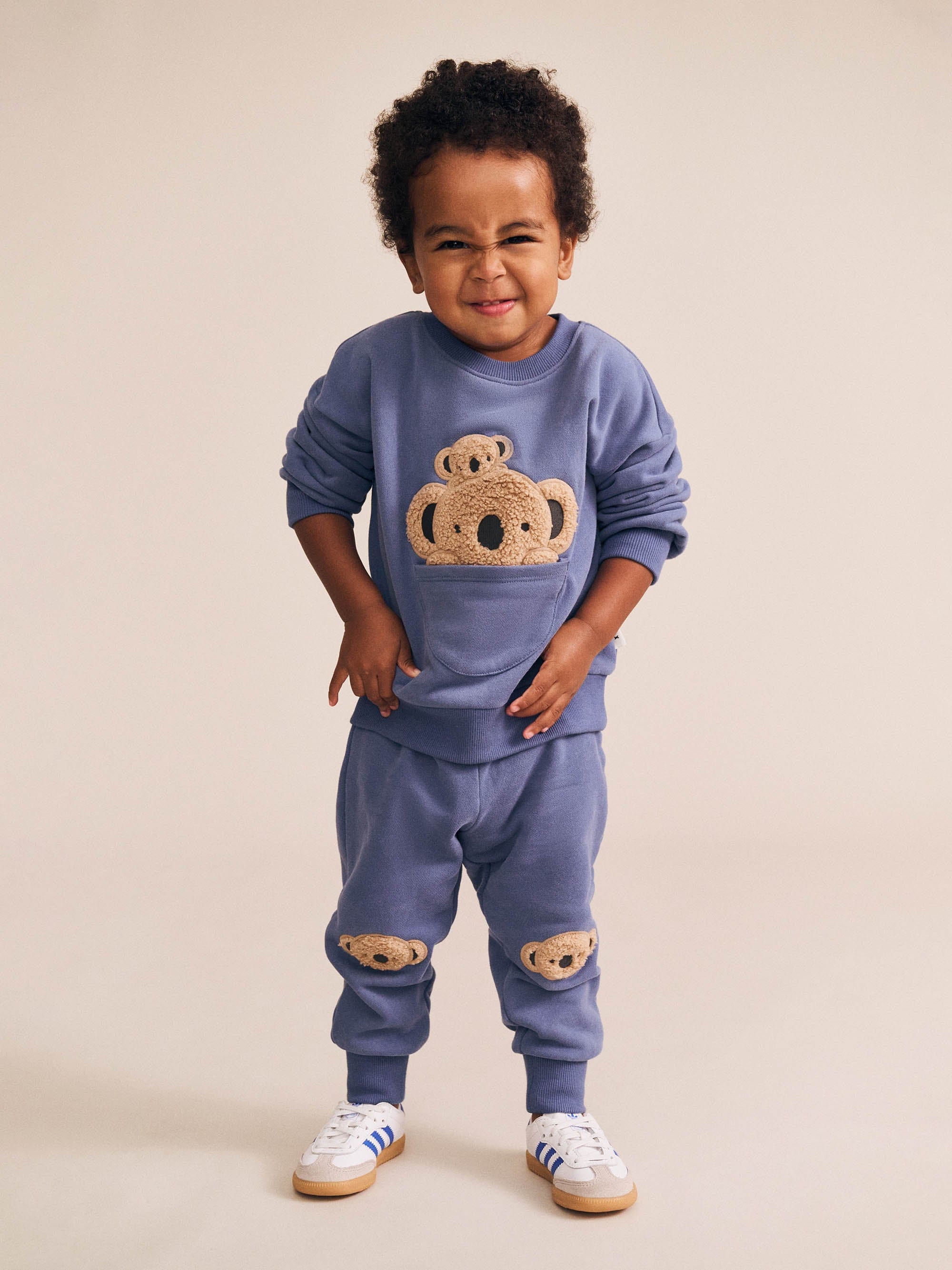 Child wearing blue Huxbaby Peekaboo Kel Pocket Sweatshirt with koala applique design in full body front view