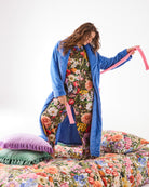 Woman wearing blue Pie In The Sky Adult Cosy Robe by Kip & Co with pink trim standing on floral bedding