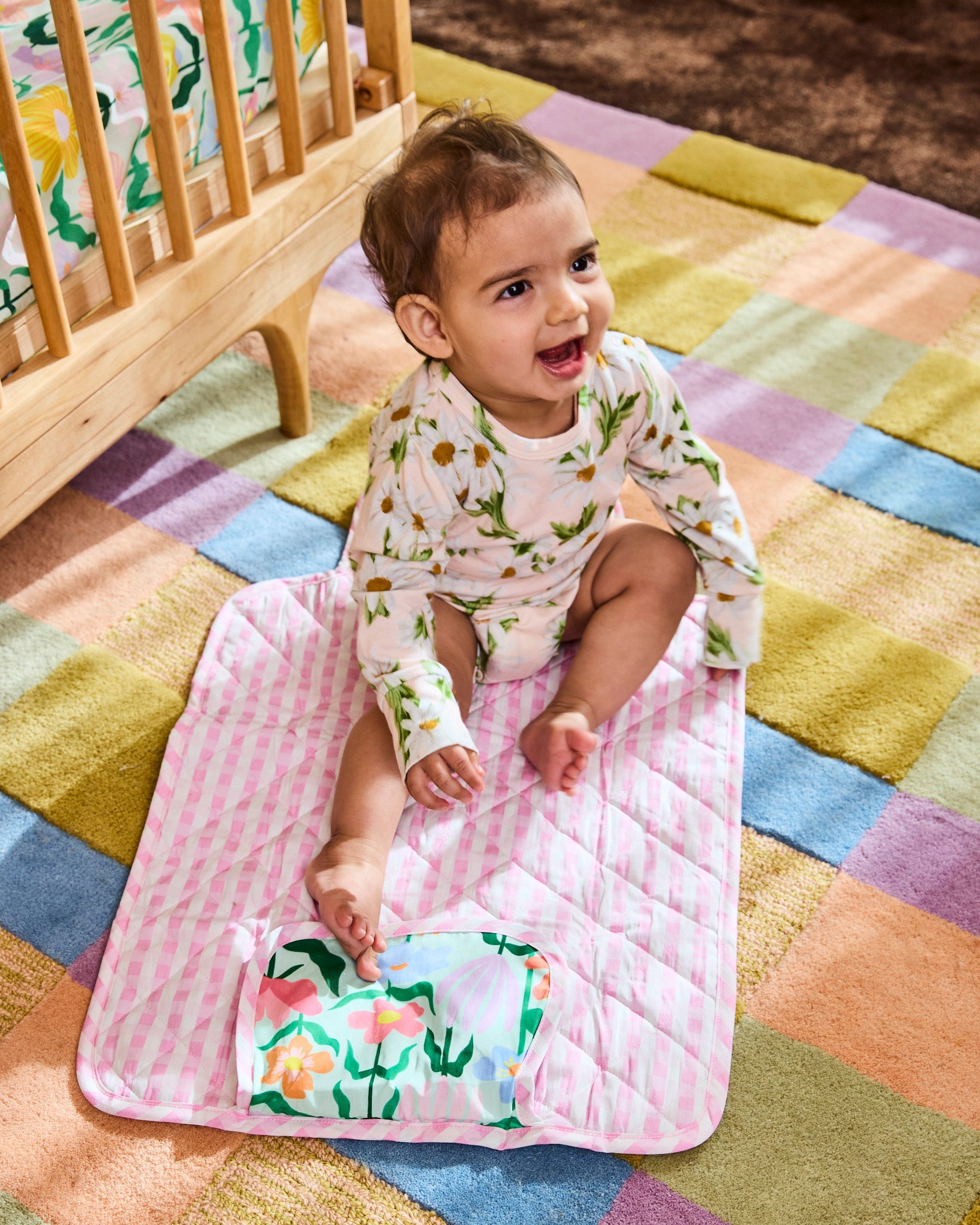 Baby sitting on pink gingham quilted change mat with Popping Flowers Green pattern detail visible at bottom corner