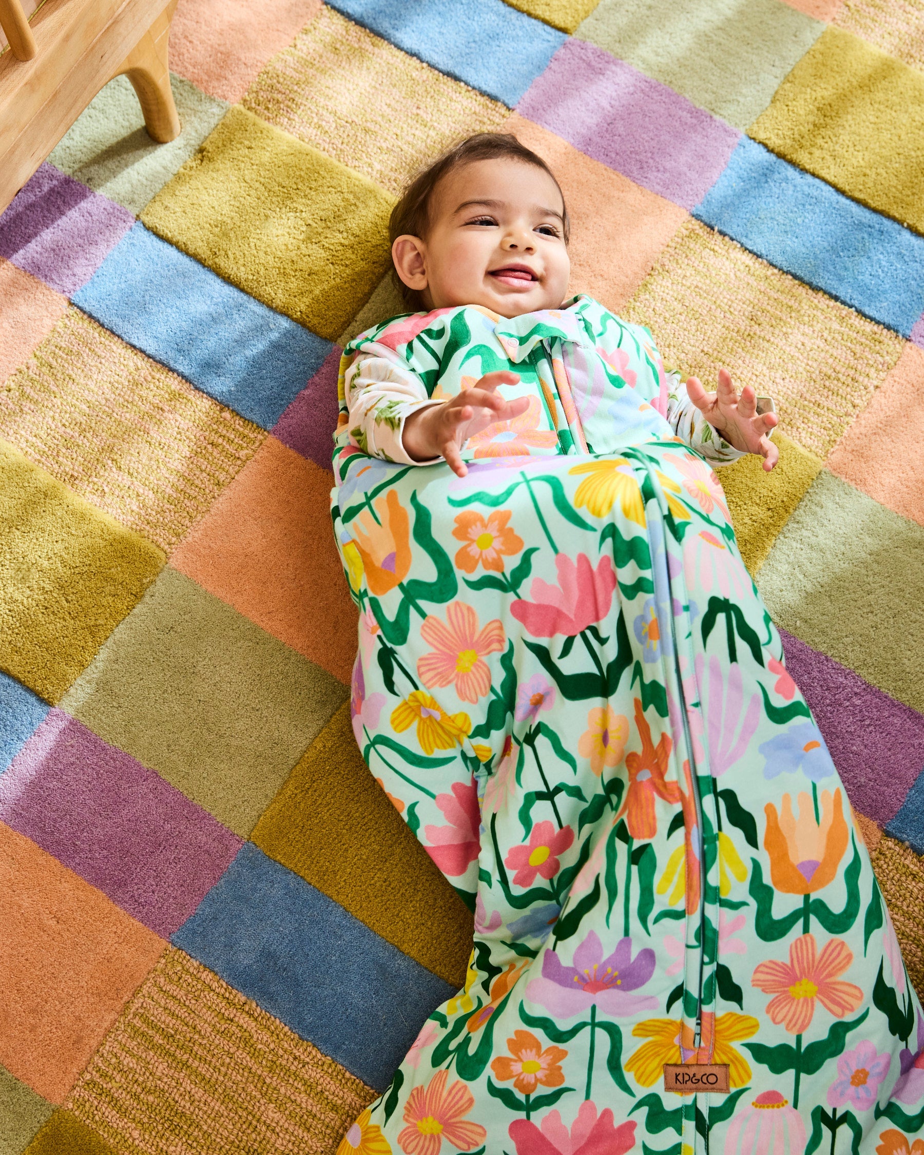 Happy baby lying in Popping Flowers Green Sleep Bag on rainbow striped rug