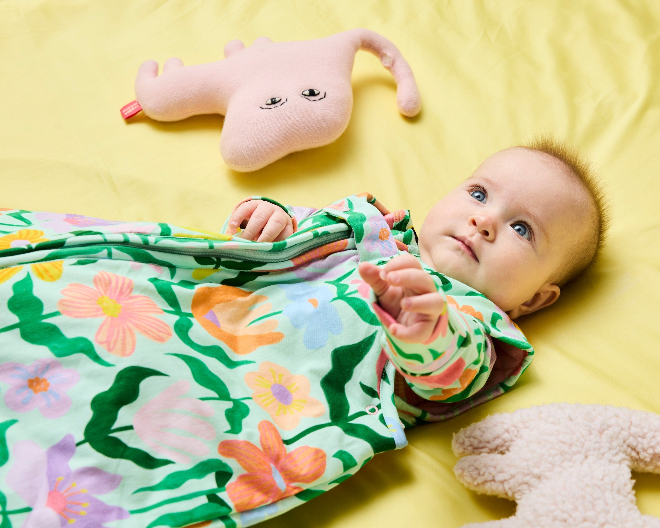 Side angle view of baby in floral sleep bag with pink plush toy on yellow surface
