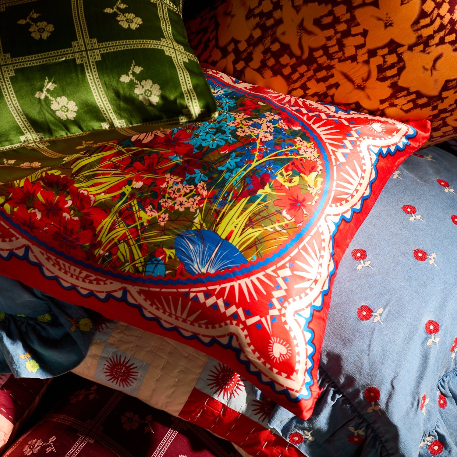Red silk pillowcase with bold floral oval print layered among colourful mixed-pattern pillowcases on a bed