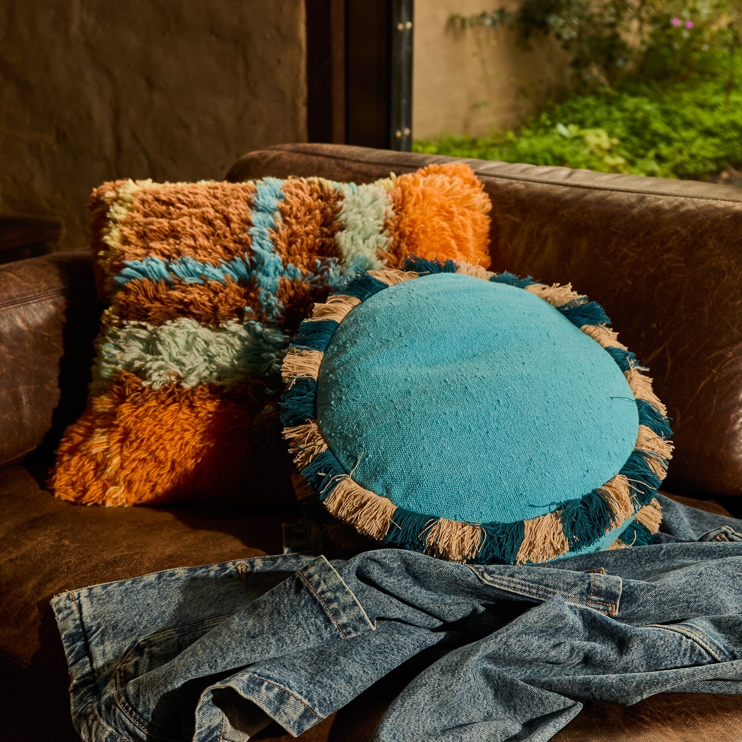Turquoise round cushion with teal and natural fringe edge resting on a leather sofa with eclectic cushions