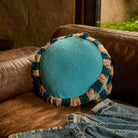 Turquoise round cushion with striped teal and natural fringe trim propped against a brown leather sofa