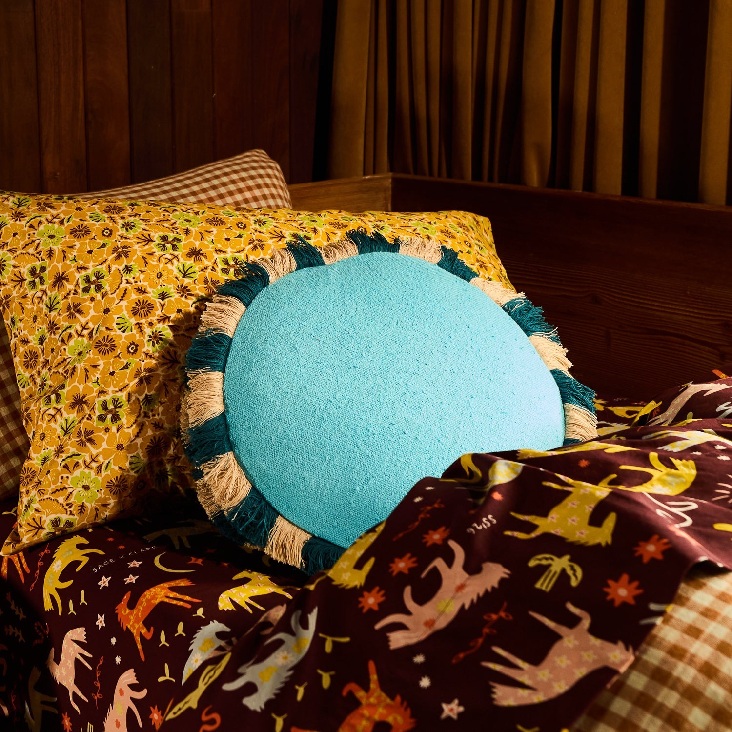 Turquoise round cushion with teal and natural fringe border placed on a patterned bed with floral pillows