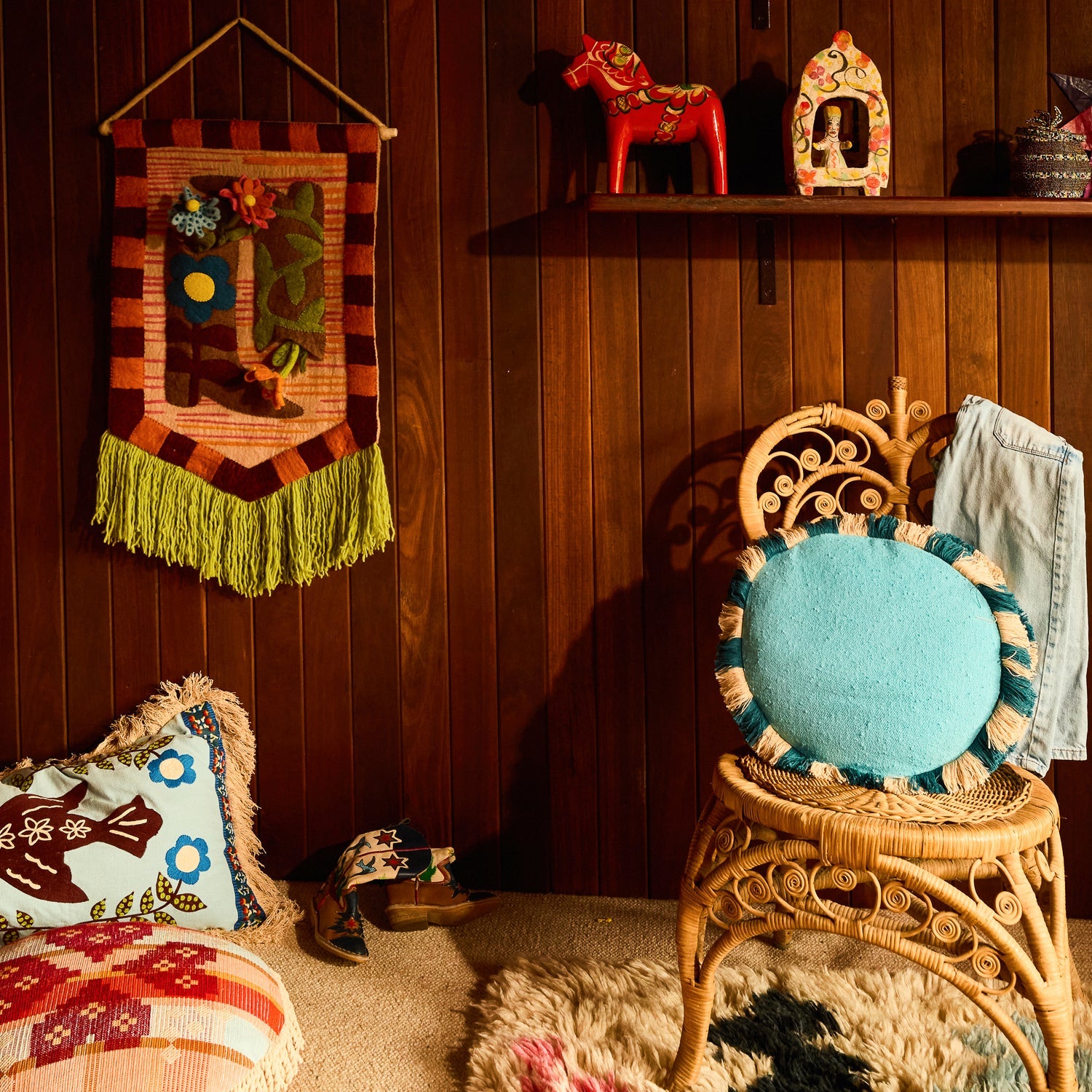 Turquoise round cushion with teal and cream fringe trim sitting on a rattan chair in a retro-styled room