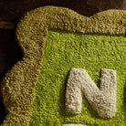 Close-up detail of the Cactus Green Nudie Rudie bath mat showing tufted texture, scalloped edge and white letter N